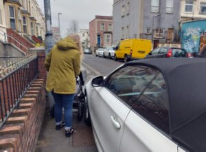 woman with pushchair squeezing through tiny gap left by car parked on pavement