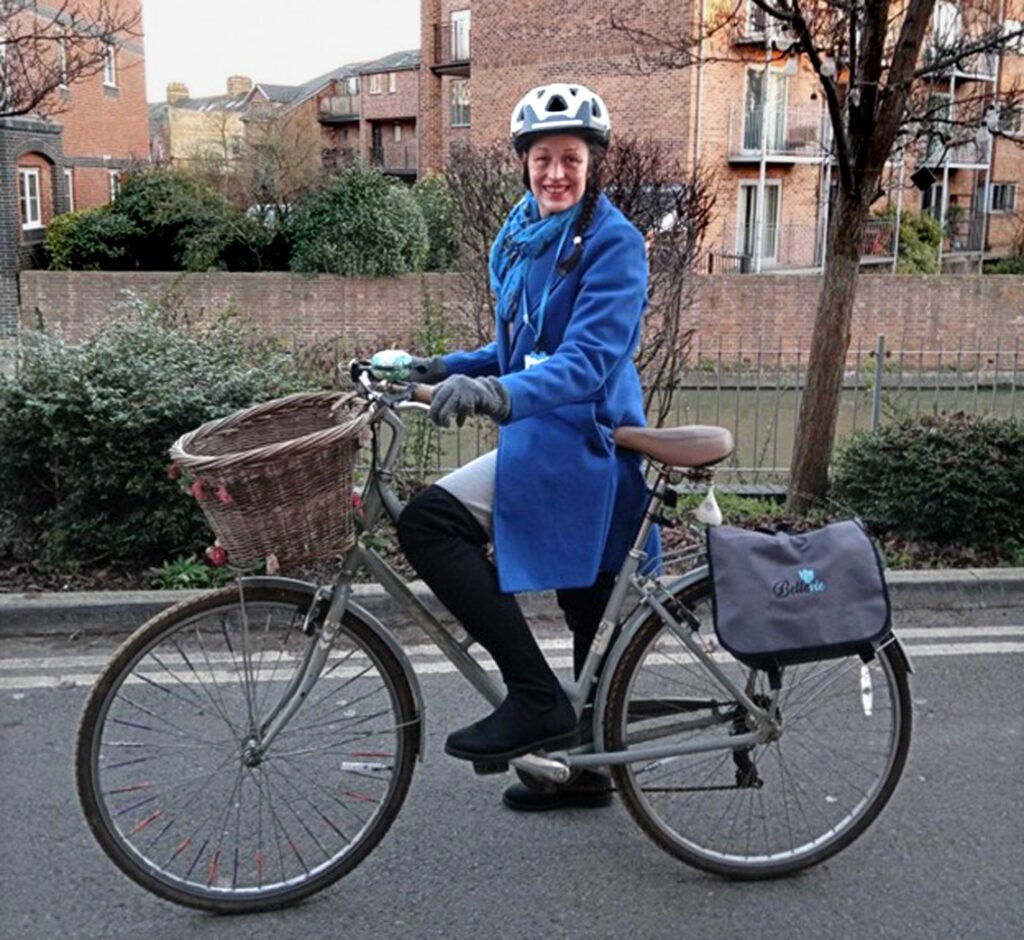 White woman in cycle helmet and blue coat posing with one foot on the pedal of sit-up-and-beg bike with basket on the front. Residential street in background
