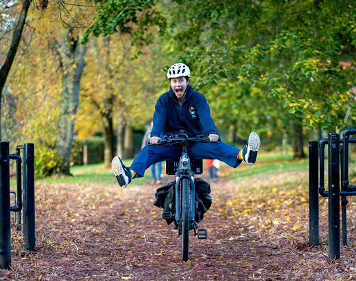 Young woman in midwife uniform and cycle helmet riding bike on a wooded path. She is shrieking with enjoyment, both feet lifted high off the pedals