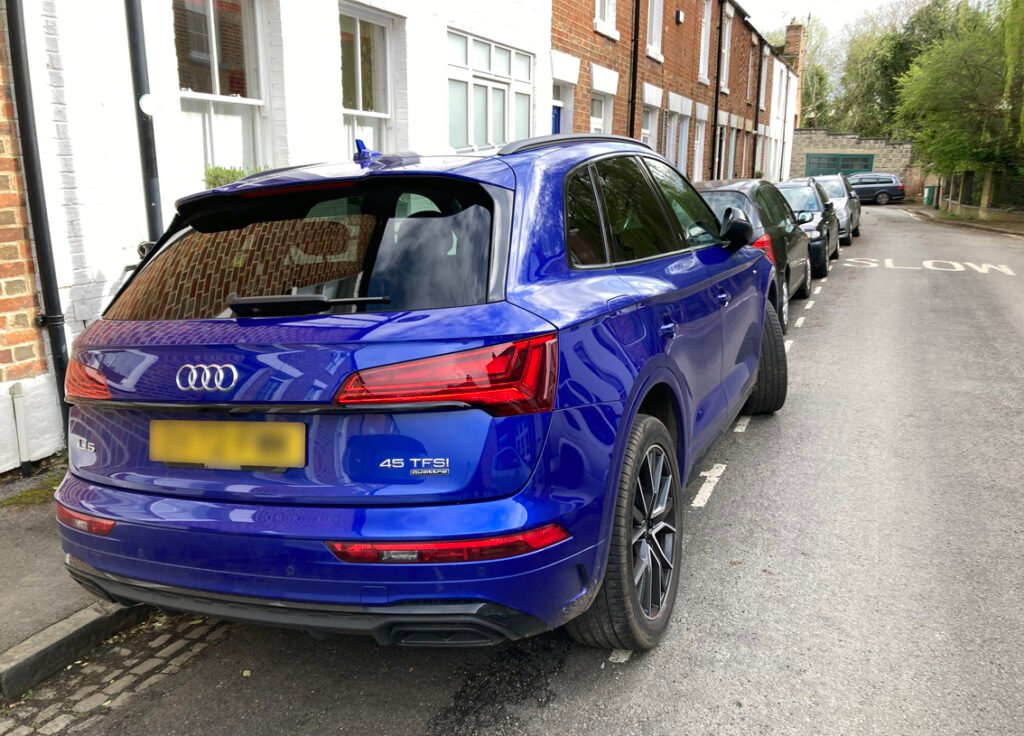 Large blue car parked at side of residential street in front of terraced houses. The car's wheels are some way over the white line for the parking bay and into the road