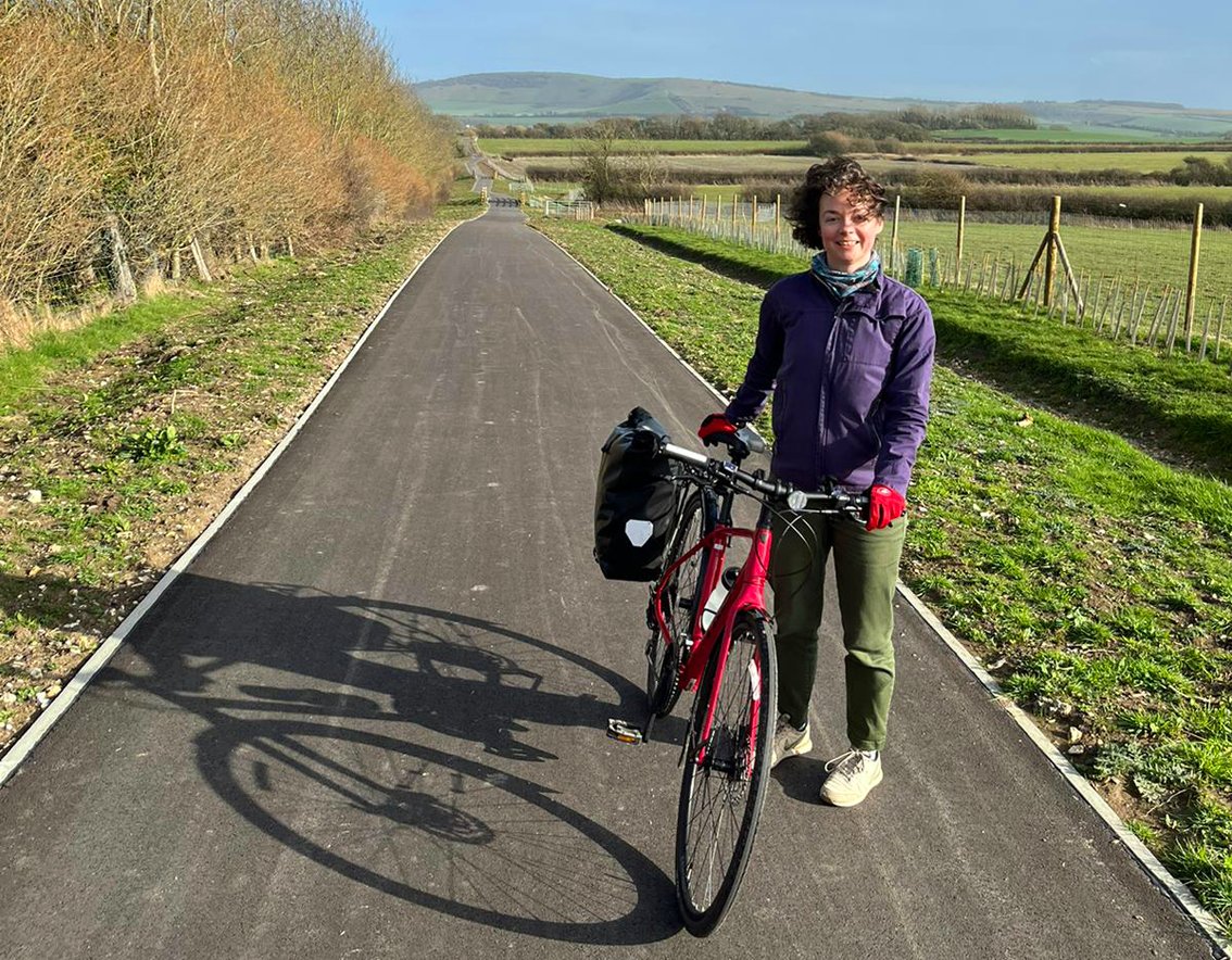 Active-looking white woman with bike standing on paved cycle path through rural landscape with blue sky in background