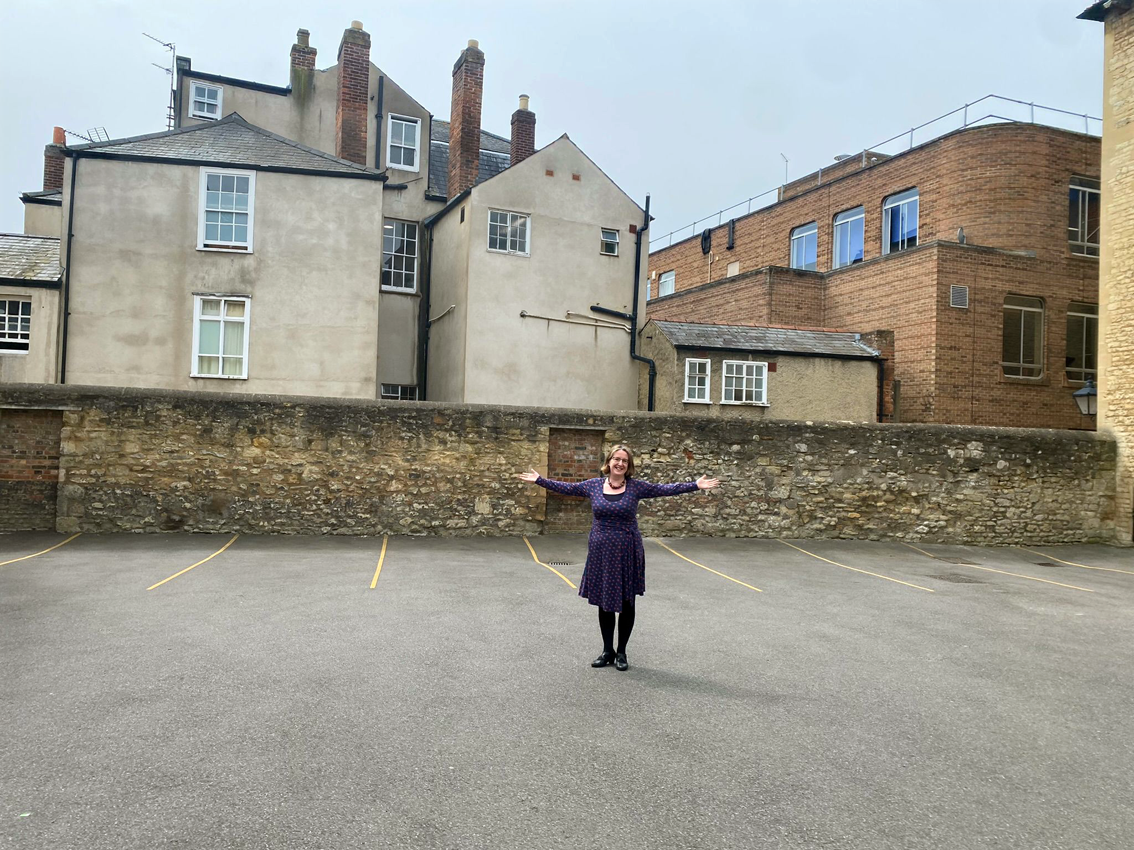 Woman standing with arms outstretched in tarmaced walled area marked out with parking spaces