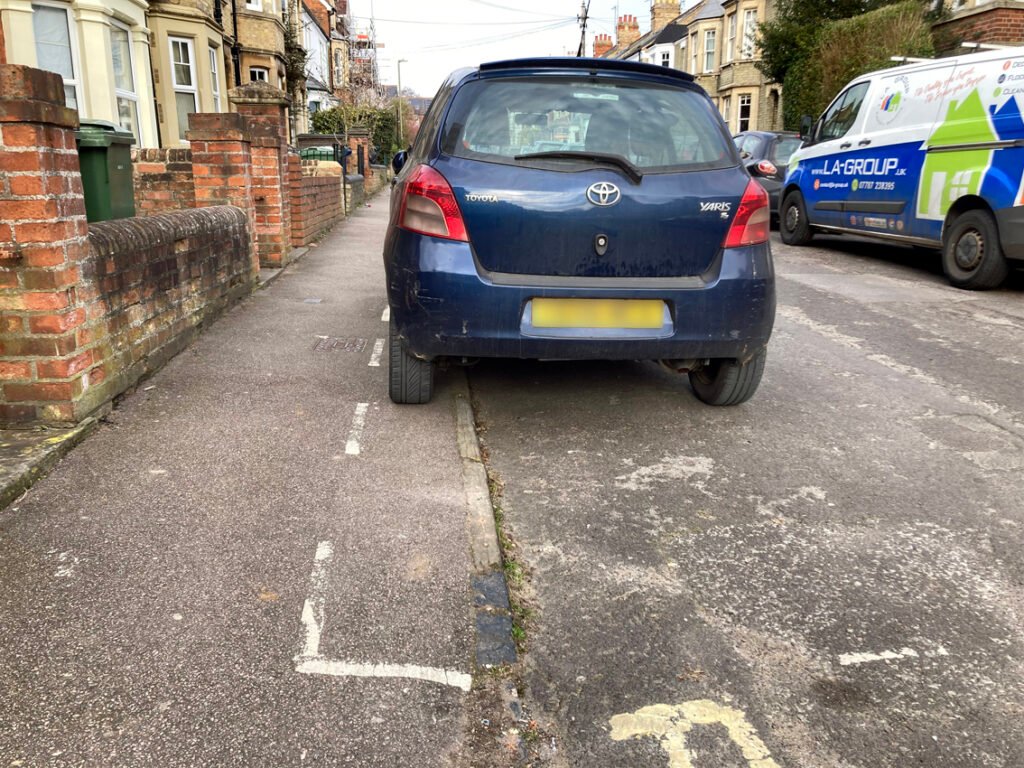 Blue car parked in parking bay that is marked out so that car obstructs half of pavement